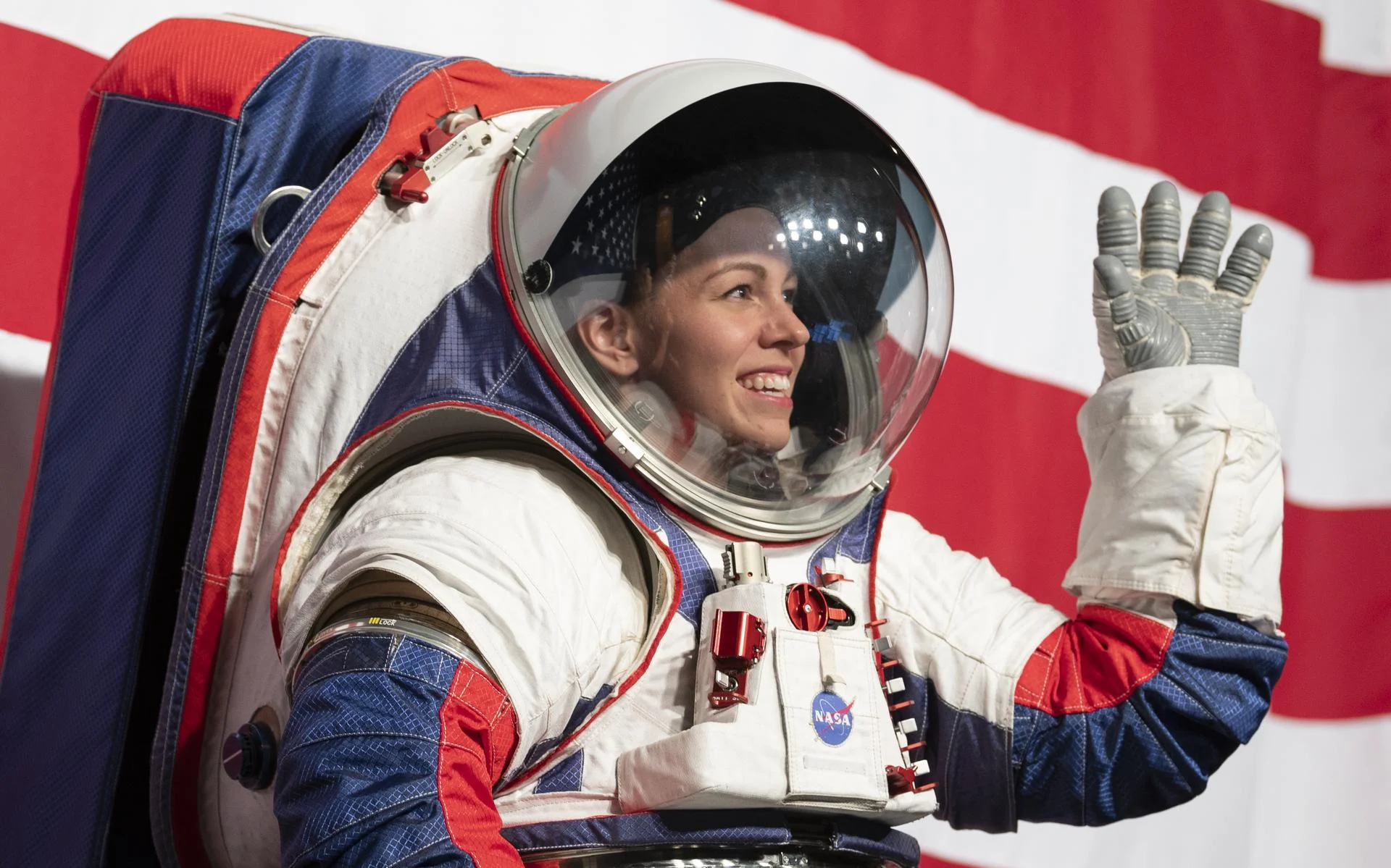 Astronaut in a red, white, and blue NASA spacesuit waving hand with an American flag backdrop.