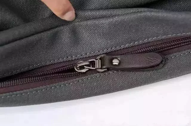 A person's finger pointing at a metallic zipper on gray fabric, showing the texture and detail of both the zipper and material.