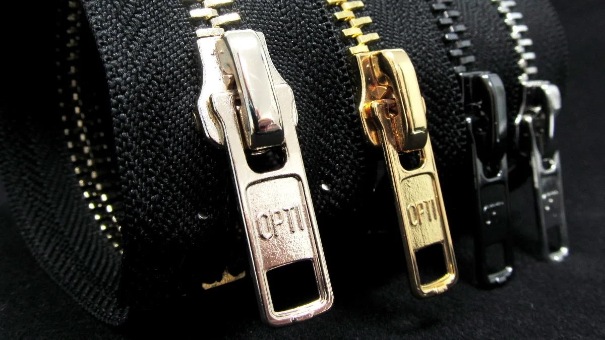 Close-up view of Opti brand zipper pulls in silver, gold, and black finishes lined on black zipper tape.