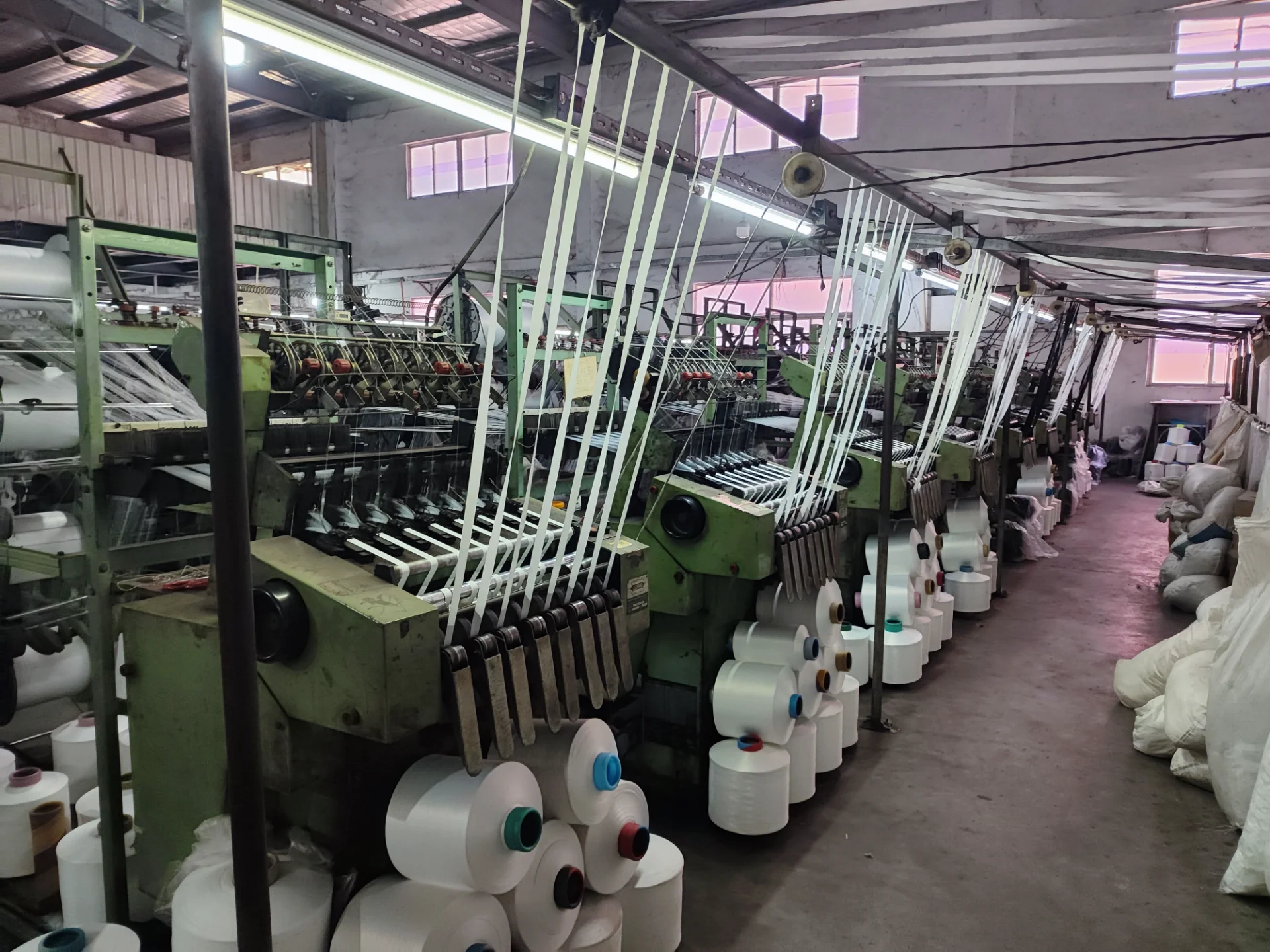 Industrial textile machines aligned in a factory with numerous rolls of threads being processed under harsh lighting, with white rolls of thread visible on the floor.