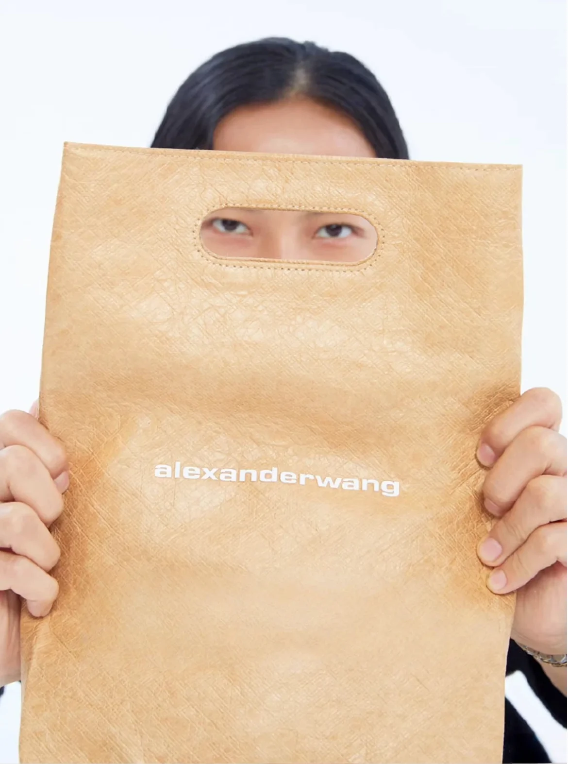 Person holding a beige Alexander Wang bag with cutout handle, covering face partially with eyes visible.