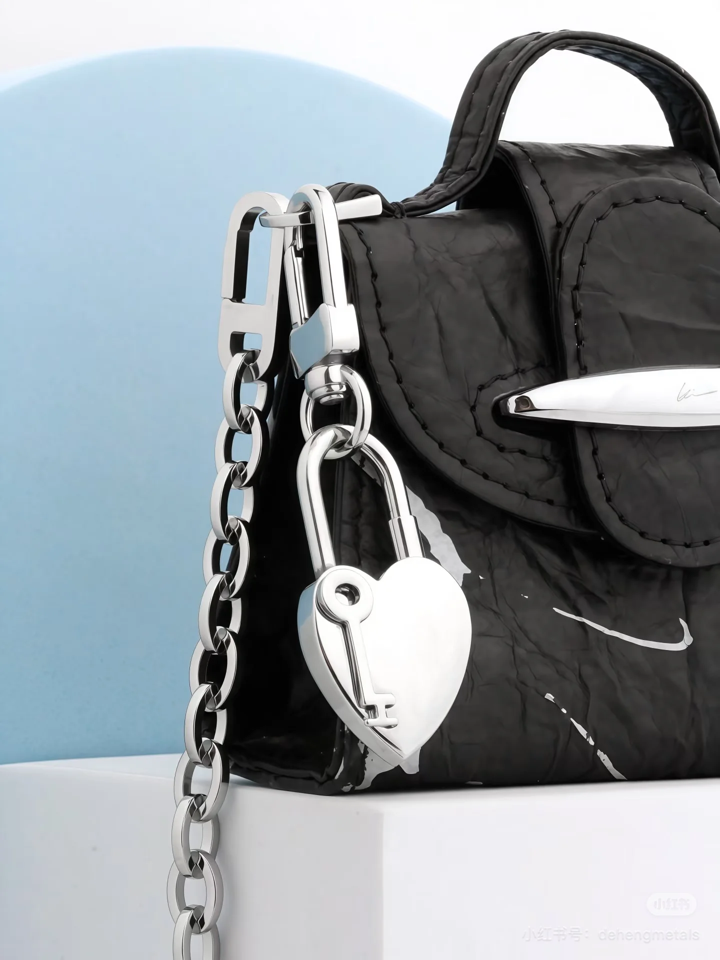 Close-up of a black textured bag with a silver chain and heart-shaped key charm against a light background.