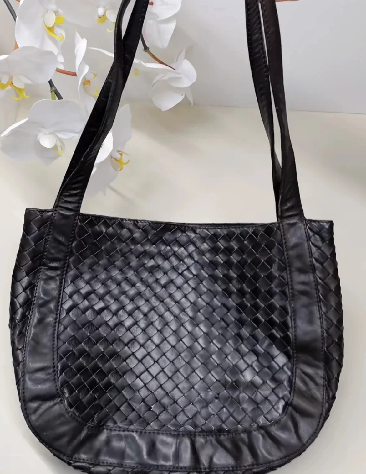 Black woven leather tote bag with long straps sitting on a light surface. White flowers in the background.