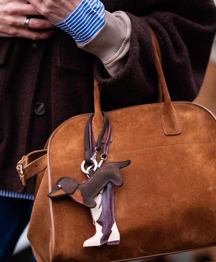 Close-up of a brown suede bag featuring a dog-shaped keychain. The bag is held by a person wearing a blue striped shirt and dark coat.