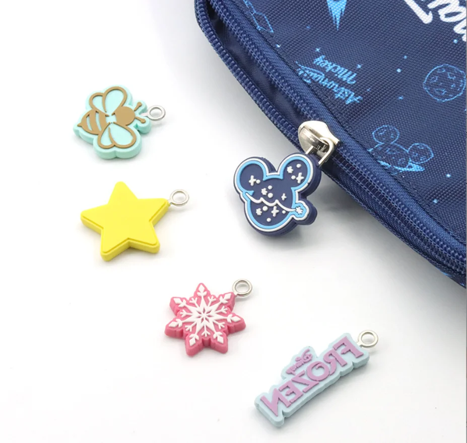 Cartoon-themed zipper pulls shaped like a bee, star, snowflake, character silhouette, and logo, placed near a blue bag.