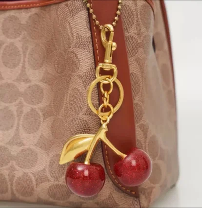 Gold and red cherry keychain attached to a brown patterned bag with leather accents.