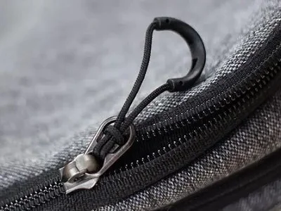 Close-up of a metal zipper with a fabric pull loop on gray textured fabric.