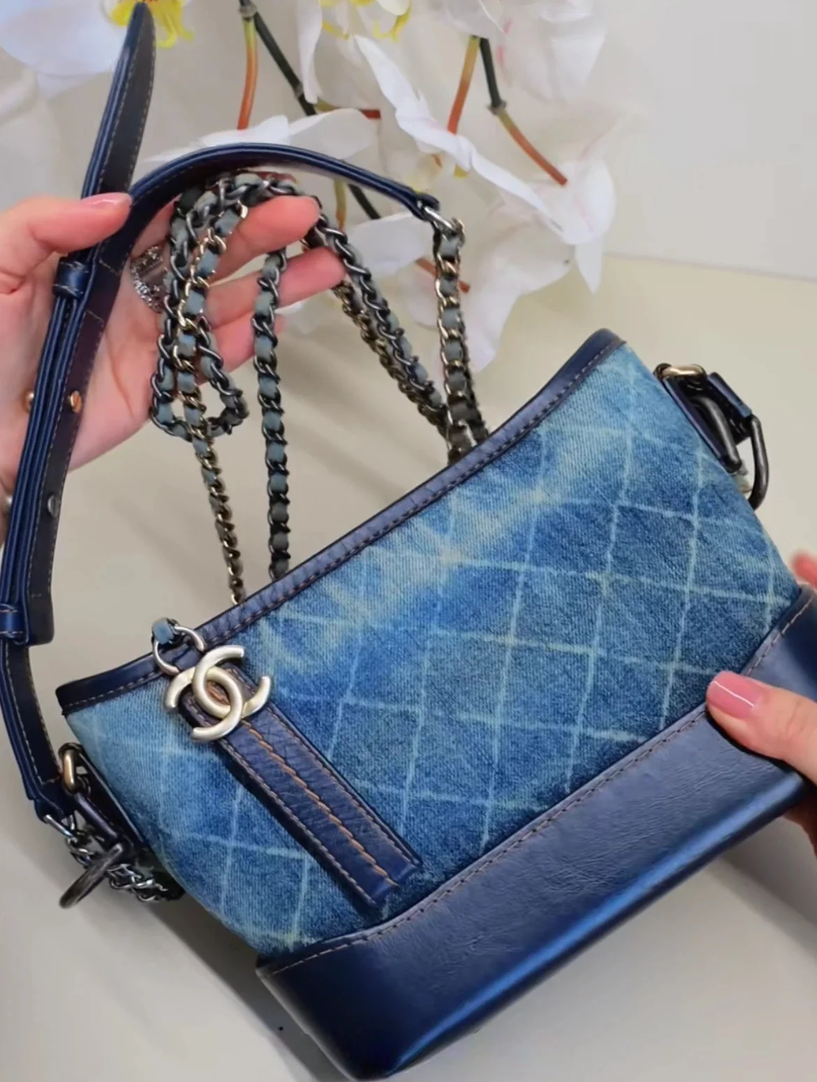 Denim shoulder bag with chain straps and leather accents held by hands, featuring a decorative logo charm.