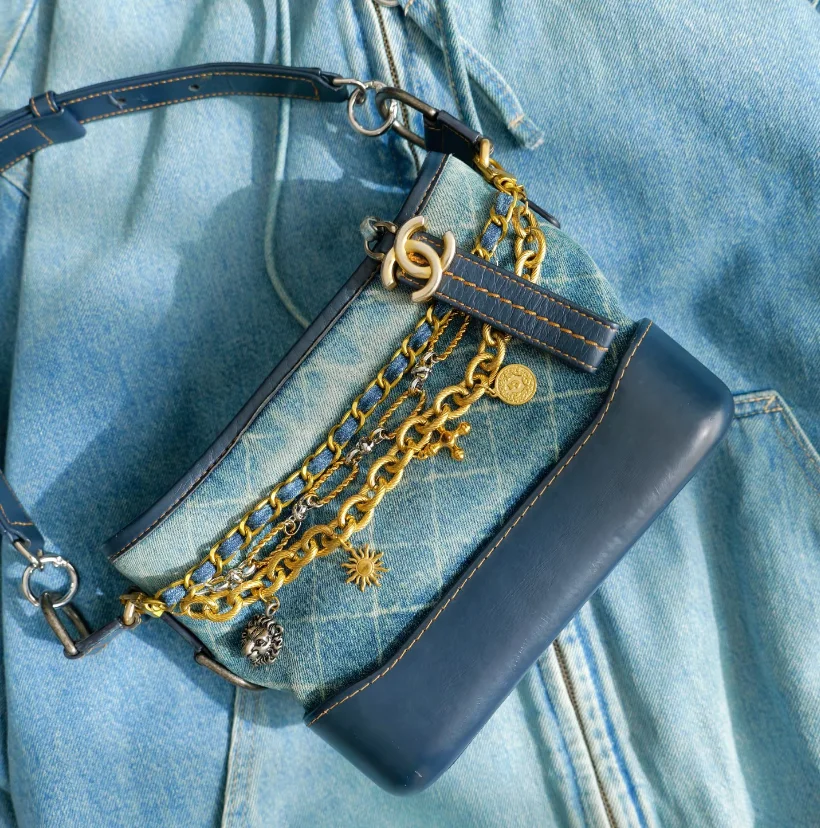Denim and leather bag adorned with gold chains, charms, and a dark blue strap, placed on a denim fabric backdrop.