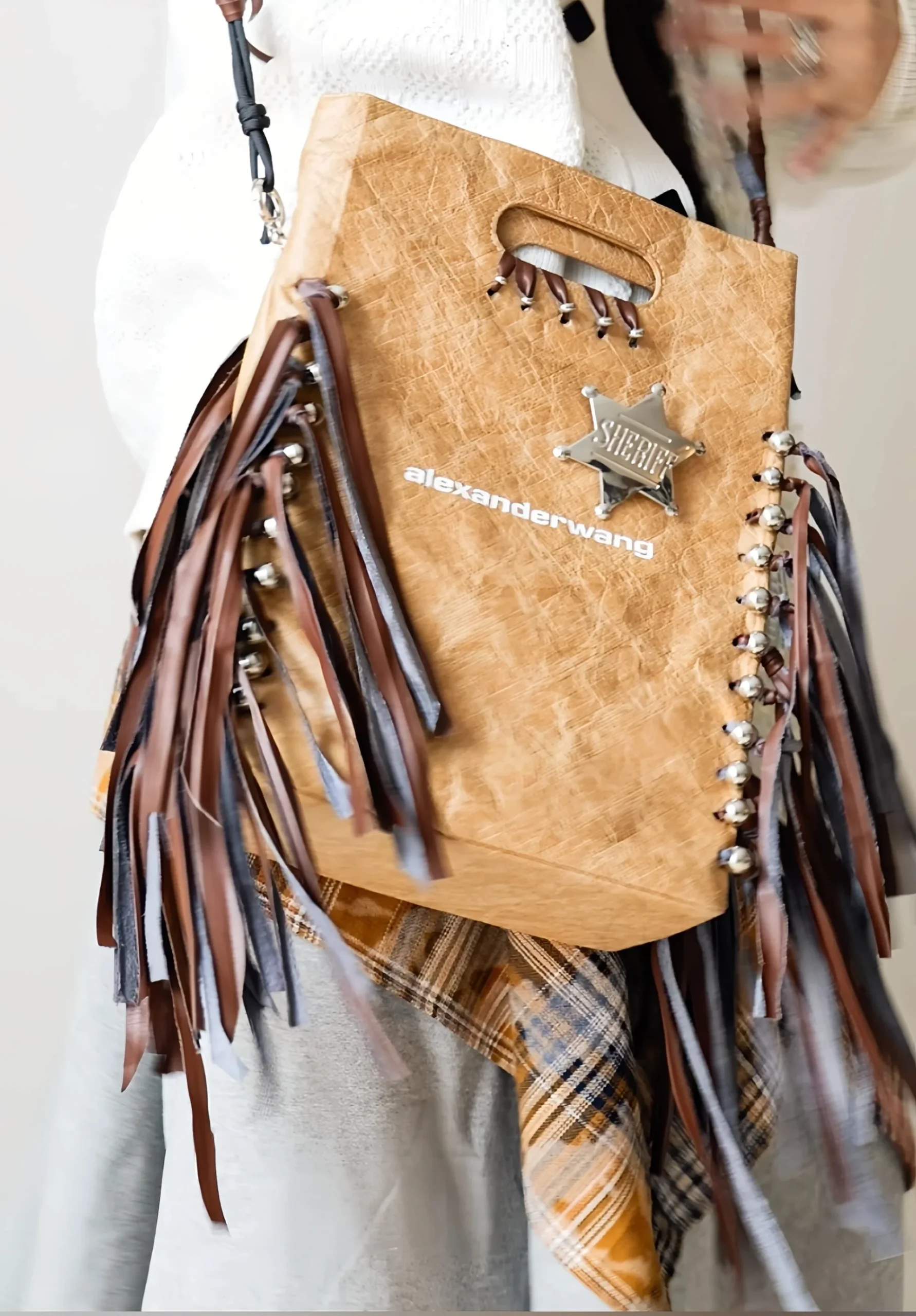 Brown fringe bag featuring a silver sheriff badge and 'alexanderwang' branding, held by a person wearing a white sweater.