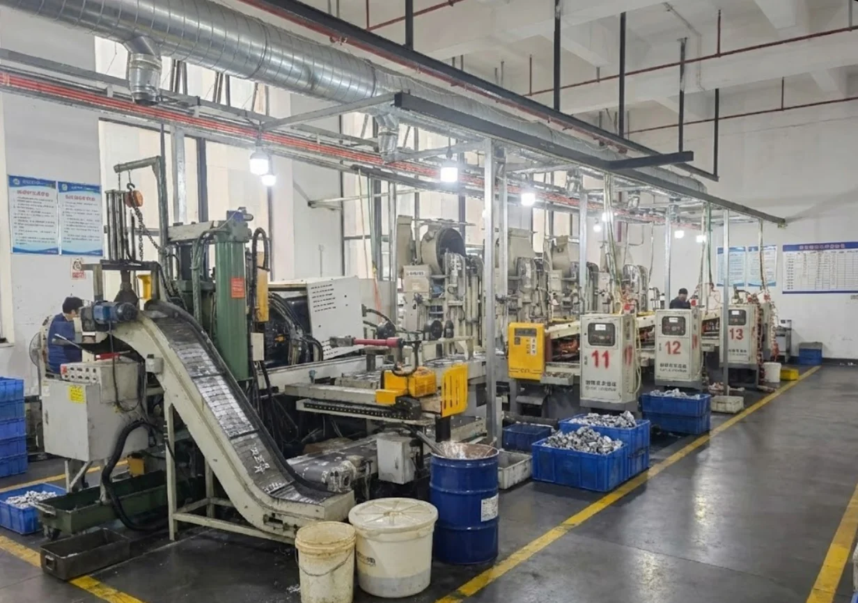 A row of industrial machines lined up in a production line within a factory space, with visible pipes and containers. Workers operate the machines.