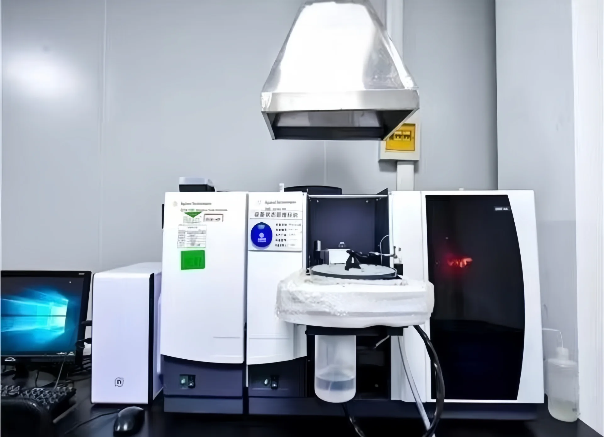 Industrial laboratory equipment including an analyzer and a computer setup on a lab bench under a ventilation hood.
