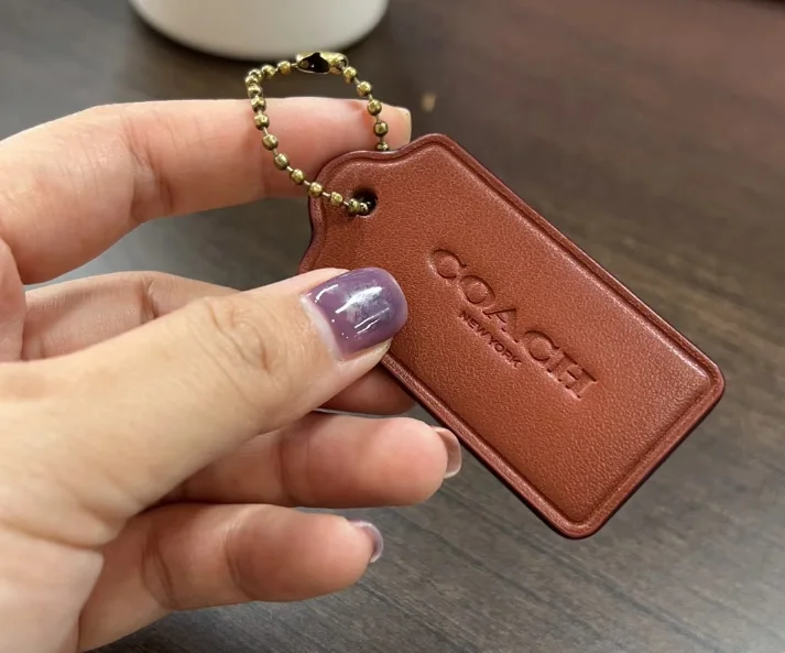 Hand holding a brown leather Coach keychain with embossed logo on a wooden table.