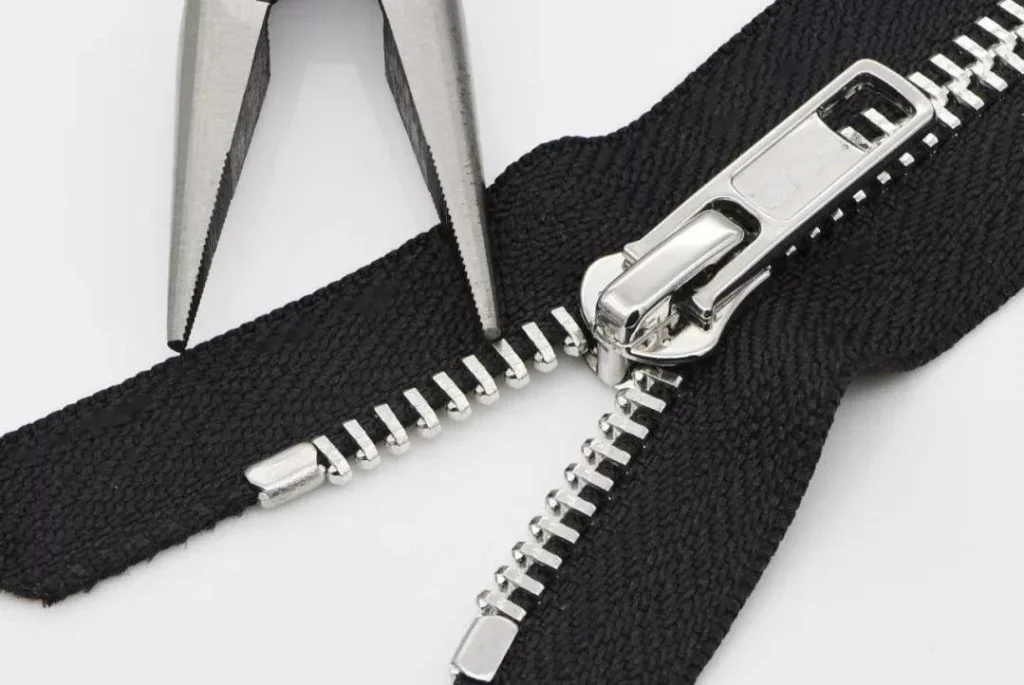 Closeup of a metal zipper with a tool holding fabric near the zipper slider on a white background.