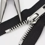 Closeup of a metal zipper with a tool holding fabric near the zipper slider on a white background.