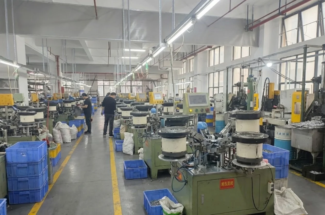 Factory floor featuring advanced textile machinery, numerous blue plastic bins, and workers managing equipment amidst bright lighting.