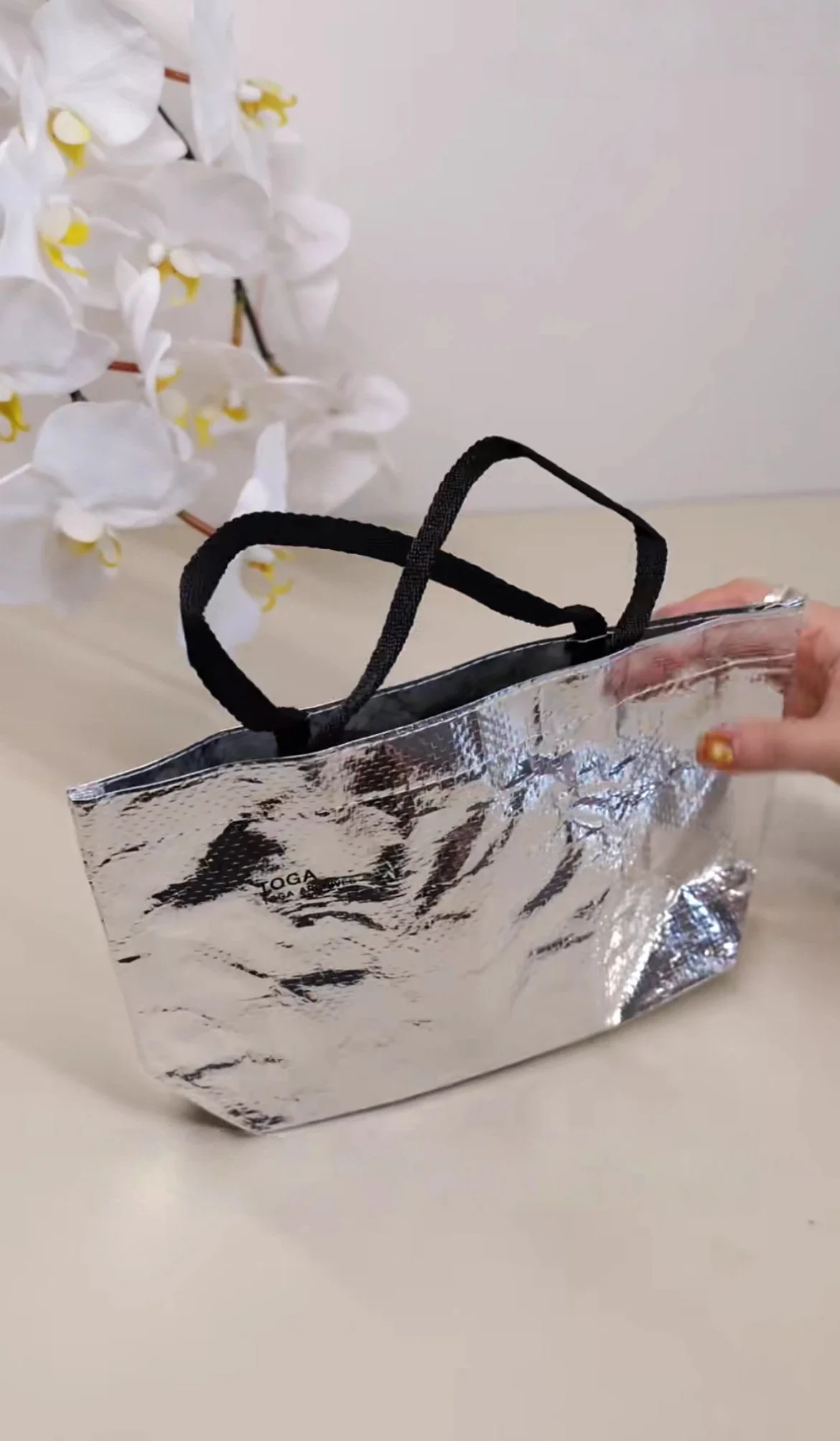 A silver tote bag with black handles is displayed on a beige surface, with a hand touching it and white orchids in the background.