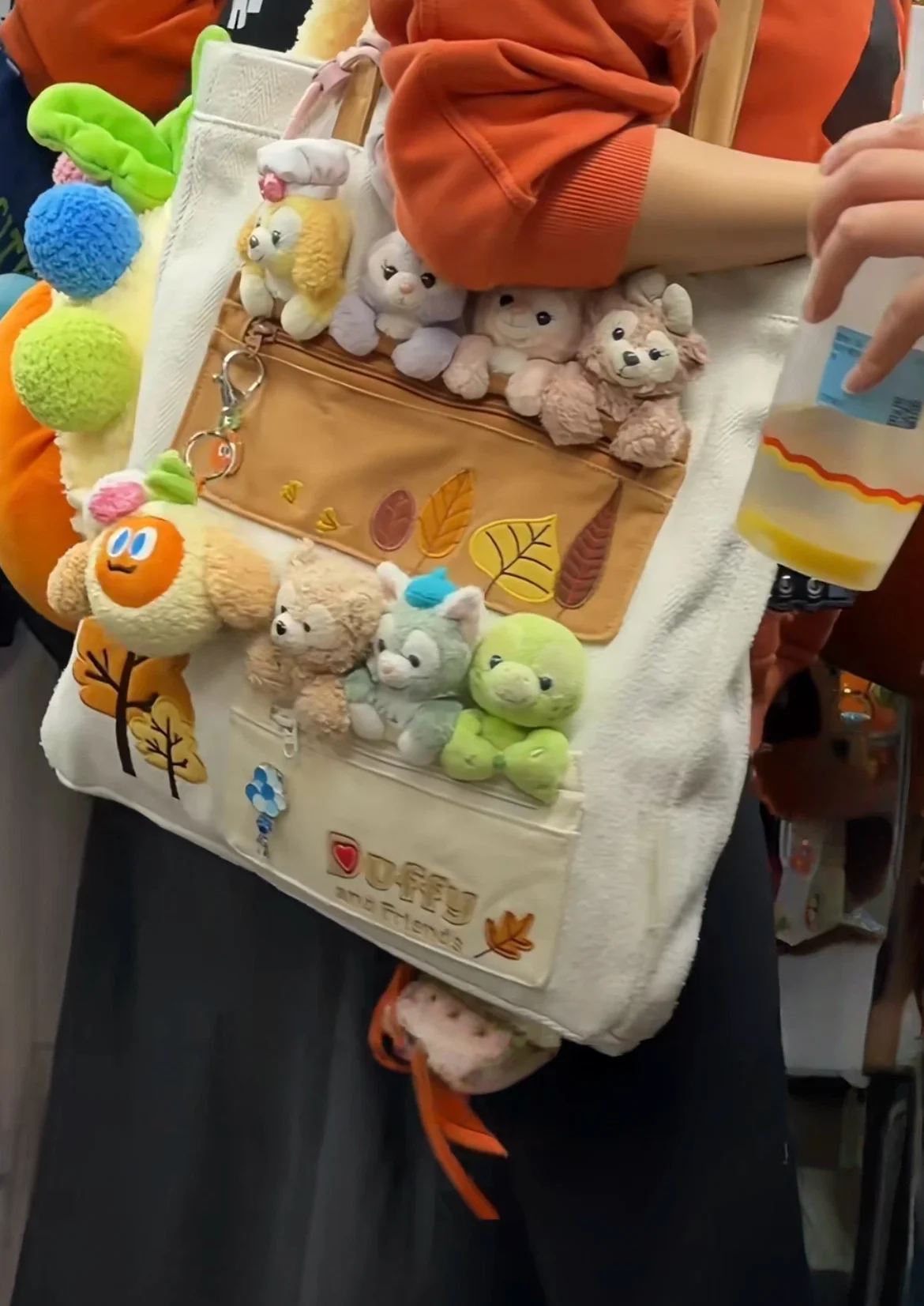 A person holds a cream-colored bag decorated with various small stuffed toys labeled 'Duffy and Friends', featuring leaf patterns and keychain attachments.