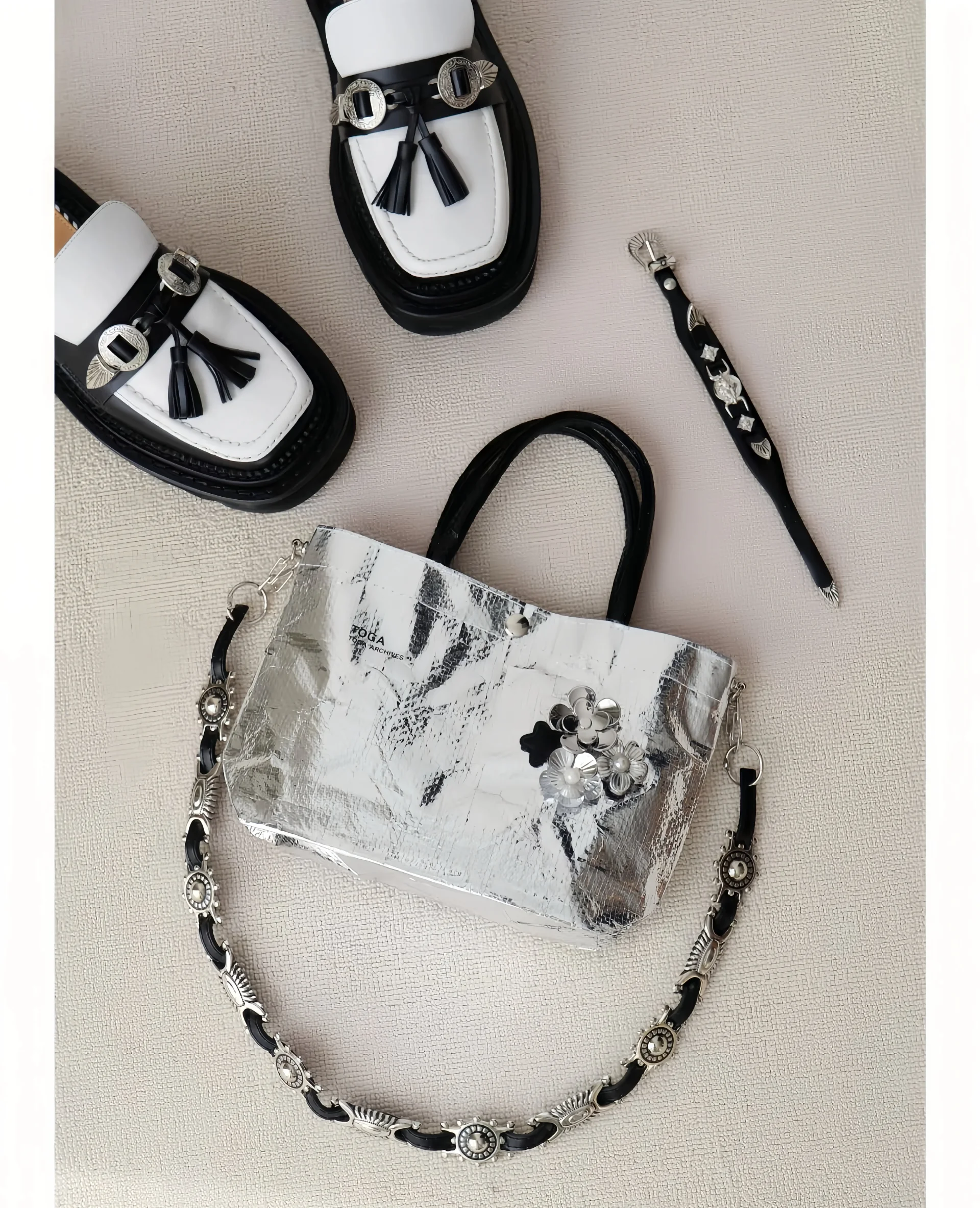 Pair of black and white loafers with tassels, a metallic silver bag with floral embellishments, and a decorative strap and accessory on a light background.