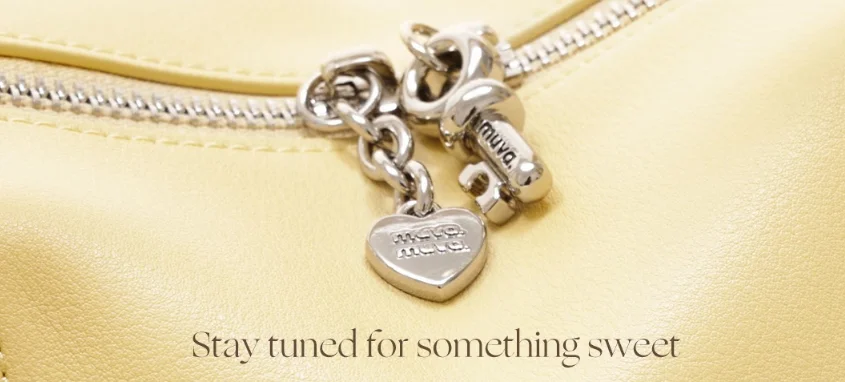 Close-up of a yellow bag with a silver heart charm and zipper, featuring engraved text and a message 'Stay tuned for something sweet.'