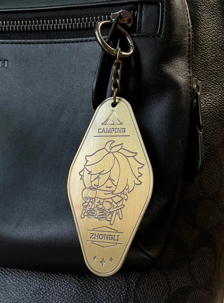 Gold metal keychain with Zhongli design attached to a black leather bag.