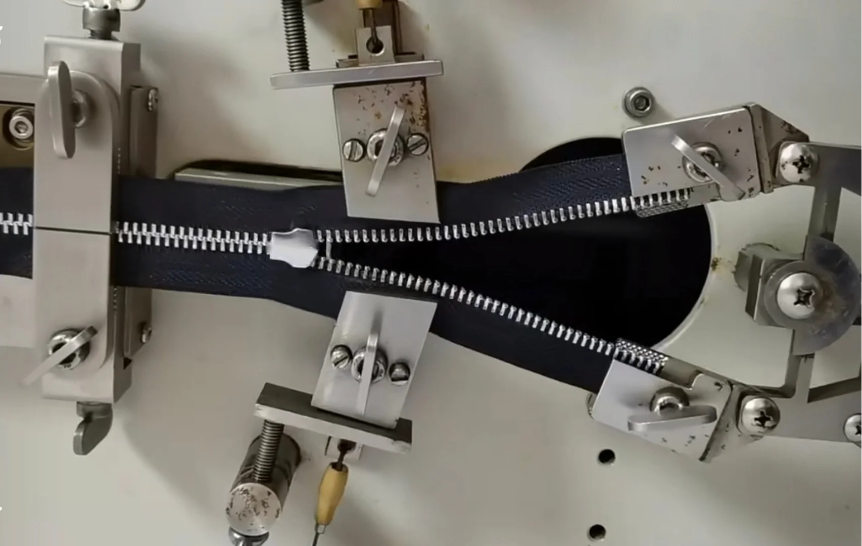 A zipper is being tested for durability on a specialized machine with various clamps and levers applying pressure.